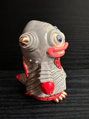 Ceramic figurine sculpture of a gray and red fish monster, with large yes and pointed ears. It has a large smile with sharp teeth.