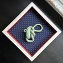 Ceramic sculpture of a green snake, seen from above. Its body is mostly coiled into itself. It sits inside of a white shadowbox with blue fabric lining the back and red fabric lining the inner borders.