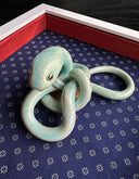Ceramic sculpture of a green snake, seen from above. Its body is mostly coiled into itself. It sits inside of a white shadowbox with blue fabric lining the back and red fabric lining the inner borders.