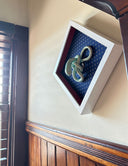 Ceramic sculpture of a green snake, seen from above. Its body is mostly coiled into itself. It sits inside of a white shadowbox with blue fabric lining the back and red fabric lining the inner borders.