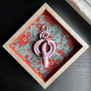 Ceramic sculpture of a reddish purple snake, seen from above. Its body is coiled into a spiral with its tail looping around itself. It sits inside of a wood shadowbox with red floral fabric lining the back and red fabric lining the inner borders.