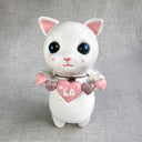 Ceramic sculpture of a white cat with large black shiny eyes and a small, cute plain expression. It holds a banner made of small ceramic hearts with the center one reading "LA". A fur collar goes around its neck.