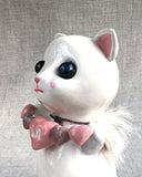Ceramic sculpture of a white cat with large black shiny eyes and a small, cute plain expression. It holds a banner made of small ceramic hearts with the center one reading "LA". A fur collar goes around its neck.