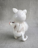 Ceramic sculpture of a white cat with large black shiny eyes and a small, cute plain expression. It holds a banner made of small ceramic hearts with the center one reading "LA". A fur collar goes around its neck.