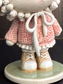Ceramic figurine of a cute anthropomorphic sheep, with large shiny eyes and curled horns. She wears a pink textured sweater and tennis shoes. Close up to show textured detail.