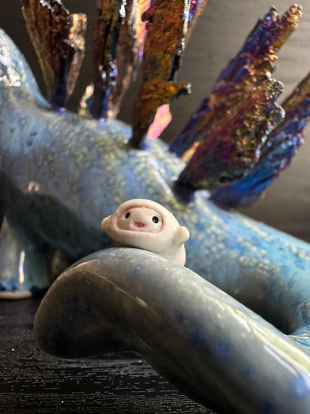 Ceramic sculpture of a blue dinosaur with sharp, colorful crystals sticking out of its back. It has a tiny character on the end of the tail.