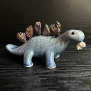 Ceramic sculpture of a blue dinosaur with sharp, colorful crystals sticking out of its back. It dangles a tiny white character out of its mouth.