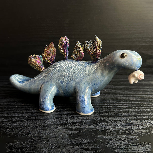 Ceramic sculpture of a blue dinosaur with sharp, colorful crystals sticking out of its back. It dangles a tiny white character out of its mouth.