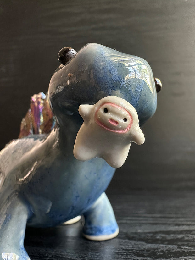 Ceramic sculpture of a blue dinosaur with sharp, colorful crystals sticking out of its back. It dangles a tiny white character out of its mouth.