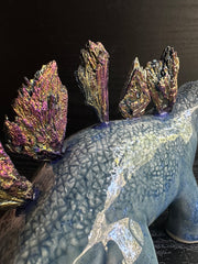 Ceramic sculpture of a blue dinosaur with sharp, colorful crystals sticking out of its back. It dangles a tiny white character out of its mouth.