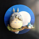 Ceramic sculpture of Totoro, positioned on a tree branch and playing a small wind instrument. Next to him are tiny Chibi Totoro, also playing instruments. Background is a circular blue sky with a giant moon.