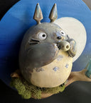 Ceramic sculpture of Totoro, positioned on a tree branch and playing a small wind instrument. Next to him are tiny Chibi Totoro, also playing instruments. Background is a circular blue sky with a giant moon.