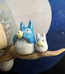 Ceramic sculpture of Totoro, positioned on a tree branch and playing a small wind instrument. Next to him are tiny Chibi Totoro, also playing instruments. Background is a circular blue sky with a giant moon.
