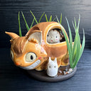 Ceramic sculpture of the Catbus from My Neighbor Totoro, with smaller proportions than usual to depict it as a kitten. Inside it is a small girl with a surprised expression. A small white chibi Totoro stands in front of the cat on a base with fake grass and small stones.