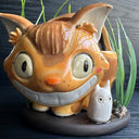 Ceramic sculpture of the Catbus from My Neighbor Totoro, with smaller proportions than usual to depict it as a kitten. Inside it is a small girl with a surprised expression. A small white chibi Totoro stands in front of the cat on a base with fake grass and small stones.