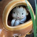 Ceramic sculpture of the Catbus from My Neighbor Totoro, with smaller proportions than usual to depict it as a kitten. Inside it is a small girl with a surprised expression. A small white chibi Totoro stands in front of the cat on a base with fake grass and small stones.