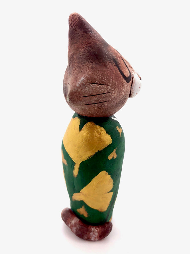 Sculpture of a large headed cartoon cat, wearing a green kimono with gold gingko leaves over it. Its eyes are closed and it has a content expression on its face.