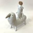 Ceramic sculpture of a bear like creature, white with smooth body, simplistic limbs and a piece of wood atop its head, riding on a bird like creature, standing on all fours with a large beak.