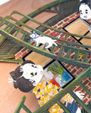Close up of an artwork of 2 characters, standing on a green fire escape on the side of a brick building, following a white cat that walks up.