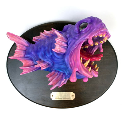 Pink and purple fish mounted to a wooden plaque. Its mouth is wide open, showing gold teeth and a red split tongue. Fish is hyper realistic in style.