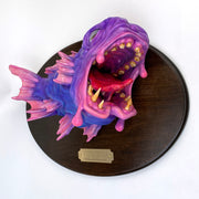 Pink and purple fish mounted to a wooden plaque. Its mouth is wide open, showing gold teeth and a red split tongue. Fish is hyper realistic in style.
