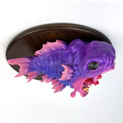 Pink and purple fish mounted to a wooden plaque. Its mouth is wide open, showing gold teeth and a red split tongue. Fish is hyper realistic in style.