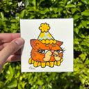 A hand holds a small square white artwork featuring a cartoon orange cat wearing a yellow party hat, with a blurred green hedge in the background.