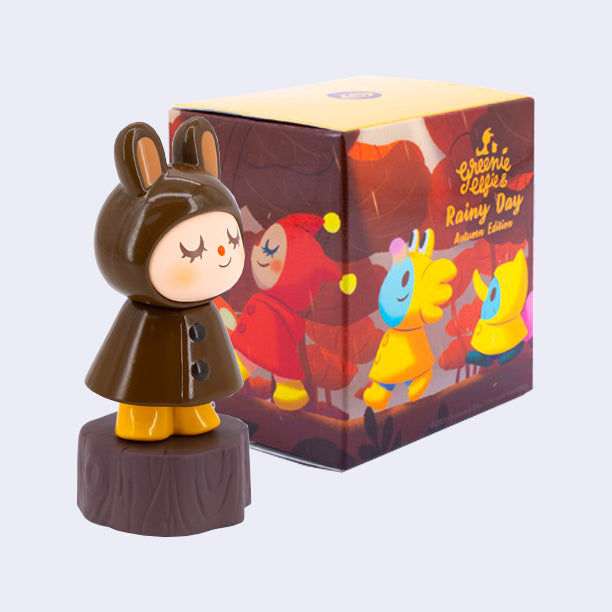 Blind box packaging and a figure of a bunny character in a brown, slick raincoat. It wears yellow boots and stands on a tree stump.