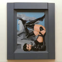 A framed painting of a stylized woman in a glossy black bodysuit and thigh-high boots, posed upside down against a gray architectural background with a light blue sky. The artwork is housed in a thick, dark gray frame and is mounted on a light beige wall.
