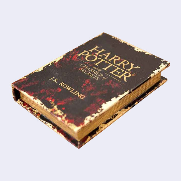 Dark brown and red hardcover book, weathered and vintage looking. Title reads "Harry Potter and the Chamber of Secrets"