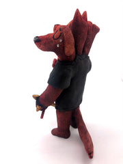Sculpture of a standing dog with 3 heads, all with their eyes closed. One looks happy, the other looks serious and the final is sad, crying tears. They wear a black shirt with a white skull, spiked bracelets and hold a red pitchfork.
