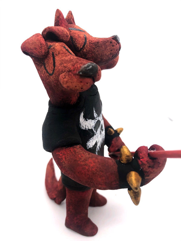 Sculpture of a standing dog with 3 heads, all with their eyes closed. One looks happy, the other looks serious and the final is sad, crying tears. They wear a black shirt with a white skull, spiked bracelets and hold a red pitchfork.