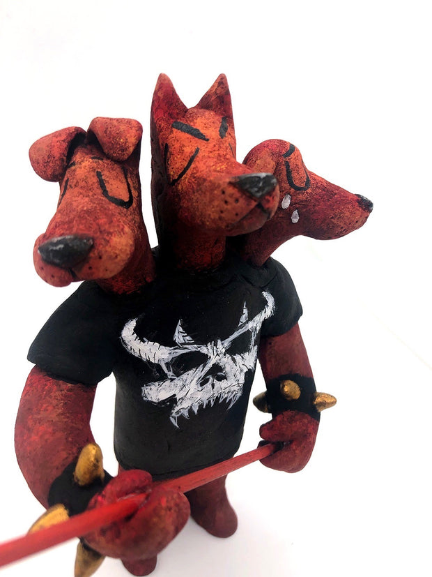 Sculpture of a standing dog with 3 heads, all with their eyes closed. One looks happy, the other looks serious and the final is sad, crying tears. They wear a black shirt with a white skull, spiked bracelets and hold a red pitchfork.