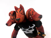 Sculpture of a standing dog with 3 heads, all with their eyes closed. One looks happy, the other looks serious and the final is sad, crying tears. They wear a black shirt with a white skull, spiked bracelets and hold a red pitchfork.