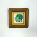 A small framed art print featuring a green cartoon creature on a white background, bordered by a yellow inner frame and a wider brown wooden frame, shown against a plain white wall.