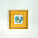 A small square artwork featuring a blue cartoon turtle character on a white background, set in a light wood frame against a plain white wall.