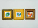 Three small framed artworks featuring cartoon creatures in olive green, mustard yellow, and brown frames, displayed side-by-side on a plain white wall.