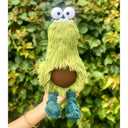 Fluffy plush sculpture of a monster shaped like an avocado, with an elongated body, large cartoon eyes and an underbite. It has a brown pit over its stomach and dangly green feet.