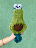 Fluffy plush sculpture of a monster shaped like an avocado, with an elongated body, large cartoon eyes and an underbite. It has a brown pit over its stomach and dangly green feet.