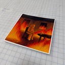 A square art print featuring a surreal illustration of a bear sitting in a burning room, photographed on a gray gridded cutting mat.