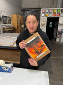 A person in a black hoodie holds up a framed artwork featuring a stylized figure in front of a fiery background, set in an art studio or workshop environment.