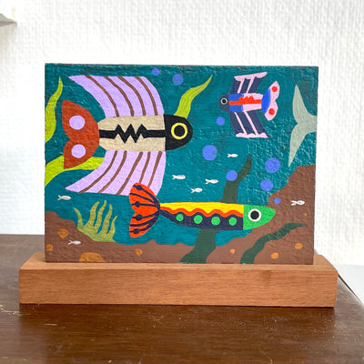 Colorful painting of 3 brightly designed fish, with different fin shapes. They swim against a teal background, with smaller fish, kelp, mud and bubbles among them.