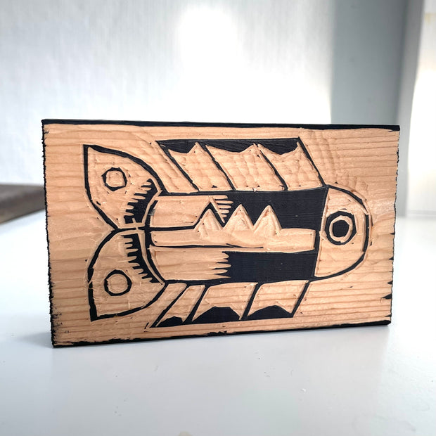 Block of wood, which has been carved into as if to make a woodblock print. The design is of a fish with straight upper and lower fins and a dual leaf like tail.