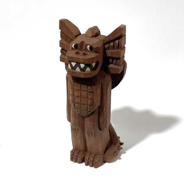 Whittled wooden sculpture of a standing dragon, with large ears, a sharp tooth mouth and a long spiked tail. It looks off to the side and has its arms at its side as it stands straight.