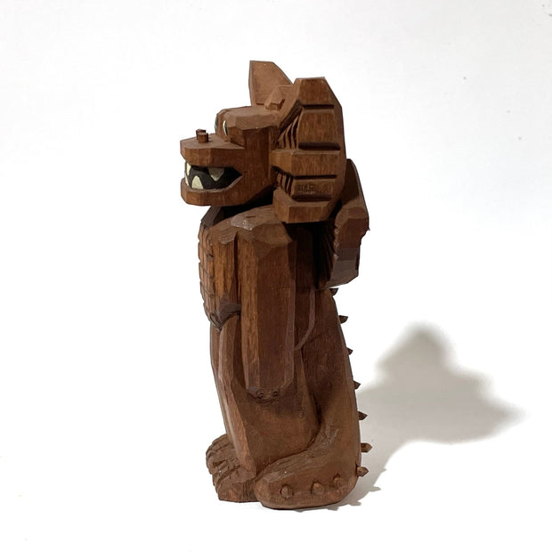 Whittled wooden sculpture of a standing dragon, with large ears, a sharp tooth mouth and a long spiked tail. It looks off to the side and has its arms at its side as it stands straight.