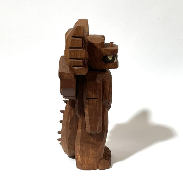 Whittled wooden sculpture of a standing dragon, with large ears, a sharp tooth mouth and a long spiked tail. It looks off to the side and has its arms at its side as it stands straight.