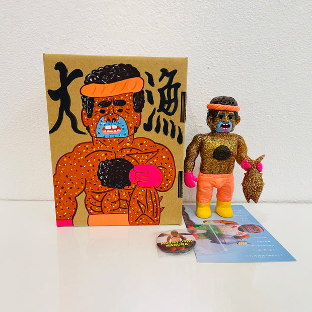 Gold glitter vinyl figure of a man, shirtless with short curly hair and chest hair. He holds a sparkly gold fish in his hands and stands besides a painted box.