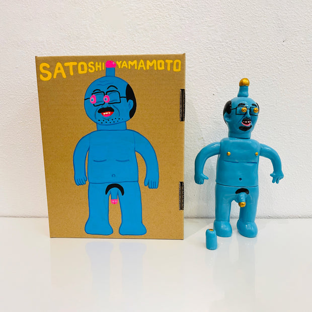 Blue painted soft vinyl figure of a nude man with glasses. He is balding with some facial hair and has a gold tipped penis atop his head. His nipples are gold and his own penis is also gold tipped. One of his hands is formed into a peace sign. Stands next to a painted box.