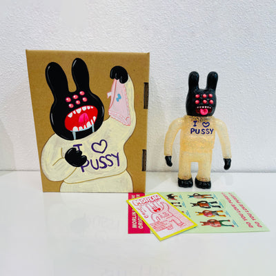 Soft vinyl figure of a black bunny monster, with 6 eyes and its tongue out. It stands wearing a glittery transparent sweatsuit that reads "I (heart) pussy". It stands next to a painted box.