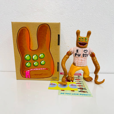 Brown painted soft vinyl figure, with 6 green eyes, a wide open mouth smile and two bunny like ears. The figure is muscular and wears a light pink shirt that says "I heart pussy" with two long tentacles as arms.